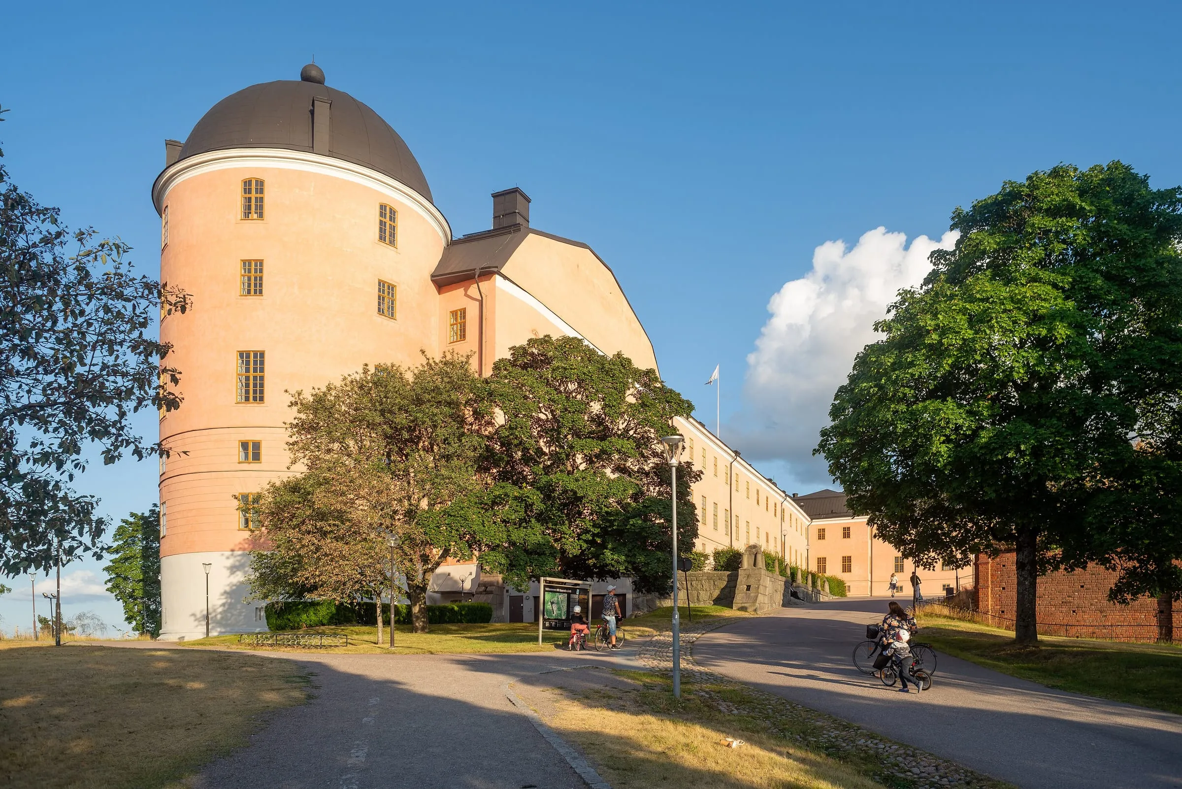 Uppsala Castle on a summer evening with its pink façade, leafy trees and a cyclist on the road in front.